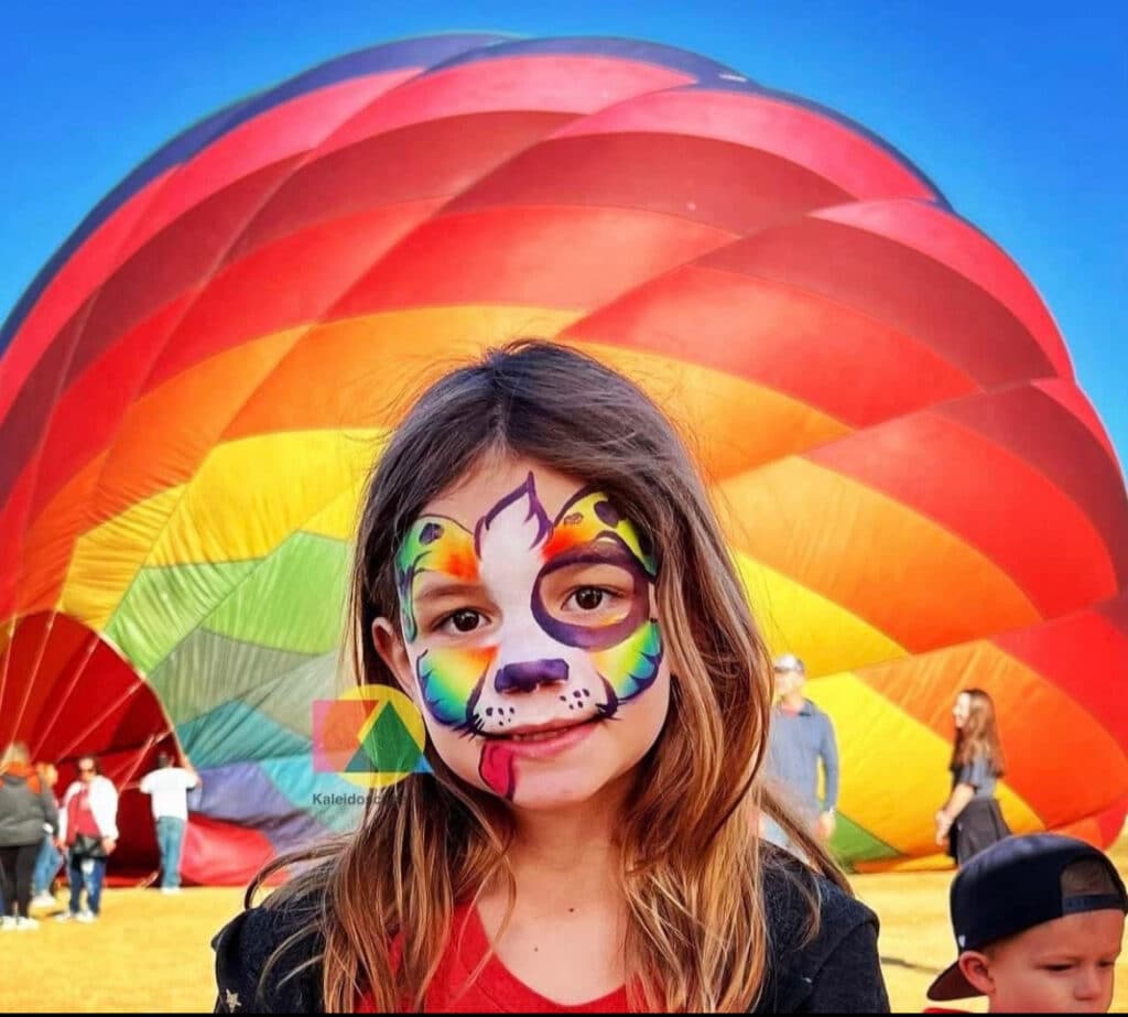 Young girl with face paint in front of a colorful hot air balloon at Balloon Festival.